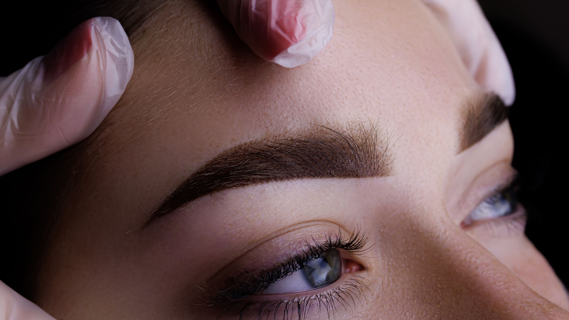 Close-up of eyebrow shaping with gloved hands and blue eye